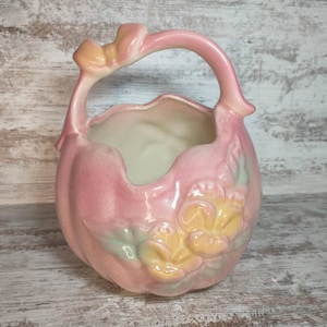 May include: A pink ceramic basket-shaped vase with a handle and floral embellishments. The vase has a light pink exterior with a white interior. A yellow and pink flower design is on the front. The handle has a bow-like detail.