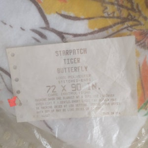 Op de afbeelding: Een wit label met zwarte tekst dat luidt: "Starpatch Tiger Butterfly 100% Polyester Genaaide uiteinden 72 x 90 inch. Wasinstructies Was &eacute;&eacute;n deken tegelijk in de machine met lauwwarm water (105 &deg;F). Fijne wasbeurt. Niet bleken. Kan in de droger worden gedroogd op lage temperatuur. Haal de deken uit de droger wanneer deze droog is of hang hem indien gewenst te drogen. 62027 Gemaakt in de VS."
