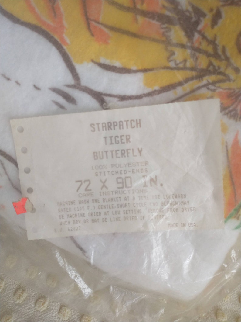 Op de afbeelding: Een wit label met zwarte tekst dat luidt: "Starpatch Tiger Butterfly 100% Polyester Genaaide uiteinden 72 x 90 inch. Wasinstructies Was &eacute;&eacute;n deken tegelijk in de machine met lauwwarm water (105 &deg;F). Fijne wasbeurt. Niet bleken. Kan in de droger worden gedroogd op lage temperatuur. Haal de deken uit de droger wanneer deze droog is of hang hem indien gewenst te drogen. 62027 Gemaakt in de VS."