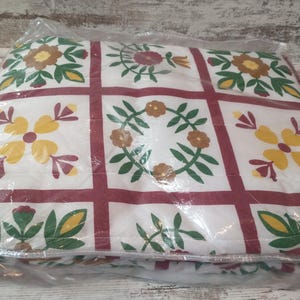 May include: A floral quilt in a clear plastic package. The quilt features a white background with a grid pattern of burgundy lines. Each square contains a different floral design in yellow, brown, and green.