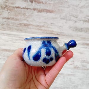 May include: A small, ceramic bowl with a handle, featuring a speckled white base and vibrant blue floral designs. The rim and handle are painted blue. This handcrafted bowl is ideal for serving small portions.