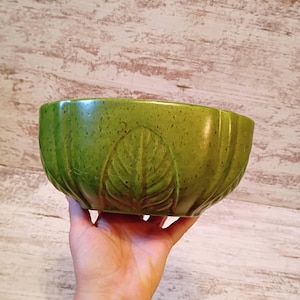 May include: A round ceramic bowl in a vibrant moss green color with a speckled finish. The exterior features a molded relief pattern of vertical leaves wrapping around the sides, creating a textured surface with deep grooves between each leaf.
