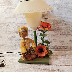 May include: Vintage table lamp featuring a cream-colored lampshade with a decorative edge. The lamp has a green stem and base, with a figurine of a person in yellow clothing. Orange flowers and green leaves add color.
