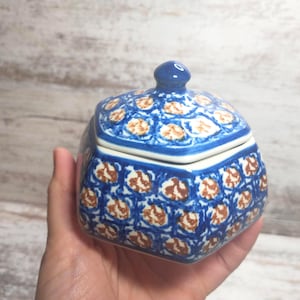 May include: A blue and white ceramic sugar bowl with a lid. The bowl has a square shape with rounded edges and is decorated with a repeating pattern of brown and white floral designs. The lid has a small blue knob.