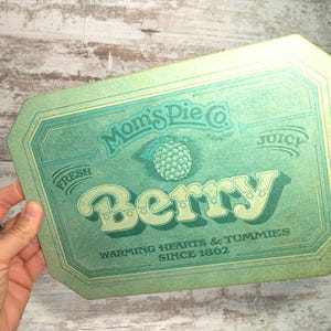 May include: A vintage-style sign with a teal and yellow color scheme. The sign reads "Mom's Pie Co. Brand" with the word "Berry" in large, decorative letters. A raspberry illustration is in the center, with the text "Warming Hearts & Tummies Since 1862".