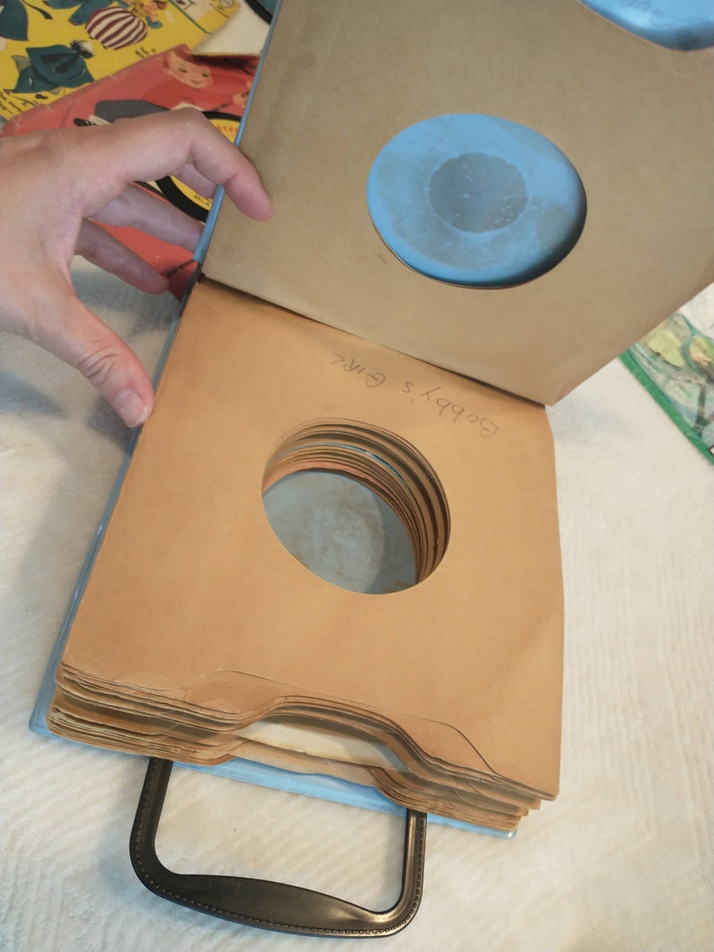 May include: A stack of brown paper record sleeves with a blue cardboard cover. The cover has a round hole and the text "Bobby's Girls" written on it.