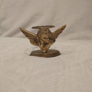 May include: A gold metal angel figurine with wings and a halo, holding a candle holder. The angel is standing on a small base.