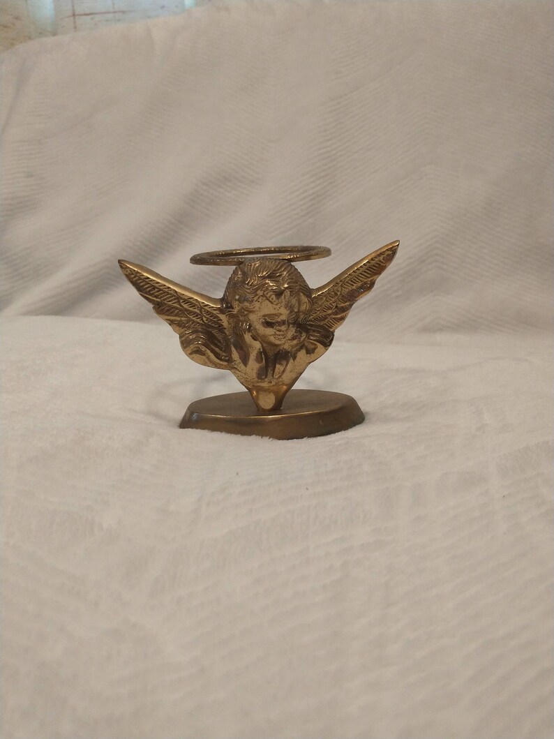 May include: A gold metal angel figurine with wings and a halo, holding a candle holder. The angel is standing on a small base.