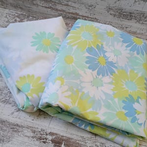 May include: Two pieces of fabric featuring a floral pattern with daisy-like flowers in shades of light blue, lime green, and white. One piece has a sparse floral print on a white background, while the other has a dense, overlapping floral design.