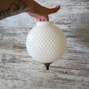 May include: A white, round, hobnail glass light fixture with a brass-colored base. The fixture is held up by a hand against a white and gray wooden background. The glass has a textured surface.