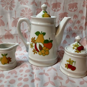 May include: A white ceramic tea set featuring a teapot, creamer, and sugar bowl. Each piece is decorated with a colorful fruit design including pears, apples, and peaches. The set is displayed on a pink floral patterned tablecloth.