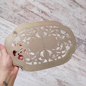 May include: An oval-shaped, metallic gold-toned decorative tray featuring an intricate, openwork filigree pattern with symmetrical scrollwork and a smooth, solid central panel. The piece has a polished finish and a scalloped, ornate outer border.