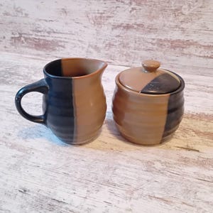 May include: A ceramic creamer and sugar bowl set. The creamer has a handle and a dark blue and tan color block design. The sugar bowl has a lid and a matching color block pattern. The set is on a light wood surface.
