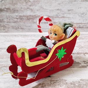 May include: A vintage-style elf figurine with blonde hair and pointed ears sits inside a red velvet sleigh. The sleigh features gold trim and a green star accent. A red and white striped candy cane is attached to the back of the sleigh.