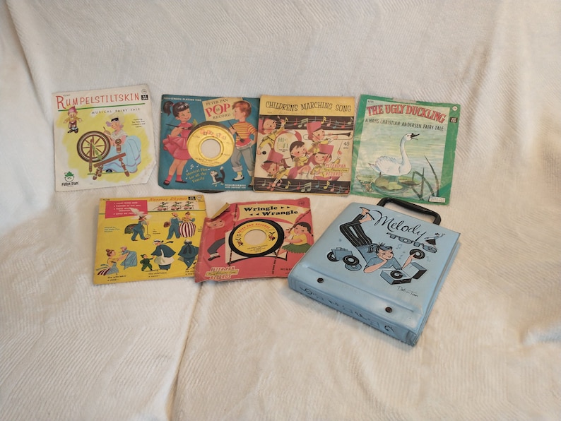 May include: A collection of vintage children's record albums and a record player case. The albums feature colourful illustrations and titles such as "Rumpelstiltskin", "Peter Pan", "Children's Marching Songs", and "The Ugly Duckling". The record player case is blue with a cartoon of a boy playing records.