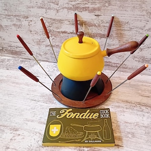 May include: A yellow fondue pot with a dark brown wooden handle and lid knob, resting on a circular wooden base with eight metal forks featuring colored tips. Beside it lies a green book titled The Fondue Cook Book by Ed Callahan.