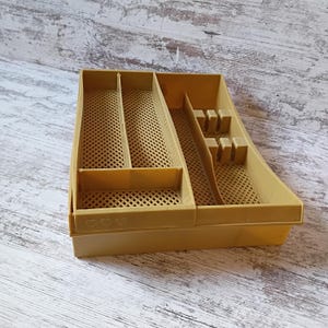 May include: A tan plastic cutlery organizer with multiple compartments. The organizer has a rectangular shape with a grid-like pattern on the bottom of each compartment. The organizer has a section for knives, forks, and spoons.