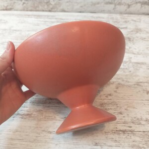 May include: A coral-colored, ceramic bowl with a rounded top and a pedestal base. The bowl is held by a hand, showcasing its smooth, matte finish. The design is simple and elegant, suitable for serving or display.