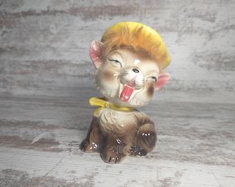 Peut inclure: Une figurine en céramique d'un chat brun et blanc portant un chapeau jaune avec de la fourrure orange. Le chat a un nez rose et sort sa langue.