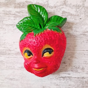 May include: A strawberry-shaped figurine with a bright red textured surface and a human-like face featuring yellow eyes and a small mouth. The top of the fruit is adorned with vibrant green leaves, and the entire piece has a glossy finish.