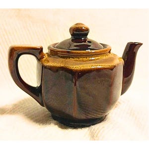 May include: A small brown and tan ceramic teapot with a lid and a handle. The teapot has a hexagonal shape.