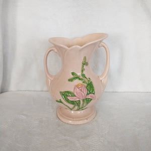 May include: A pink ceramic vase with two handles and a floral design. The vase features a pink flower with green leaves.