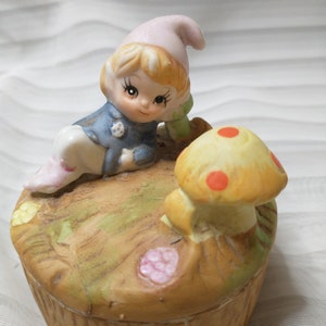 May include: A ceramic trinket box featuring a whimsical gnome figure with a pink hat and blue jacket, resting on a mushroom-shaped lid. The lid has a mushroom with orange spots. The box is painted in earthy tones.