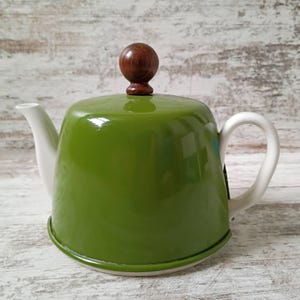 May include: A glossy olive green teapot featuring a white spout and a white handle. The lid is topped with a round, dark brown wooden knob. The teapot has a wide, rounded body and a smooth, reflective finish on its exterior surface.