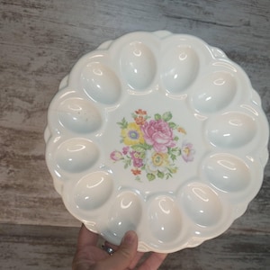 May include: White ceramic egg serving platter with scalloped edges and a floral center design. The platter has 12 egg-shaped indentations. The floral design features pink, yellow, and purple flowers.