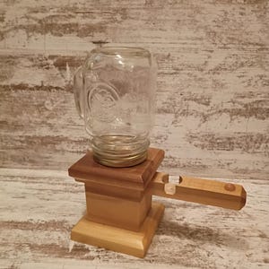 May include: A clear glass mason jar mug with a handle is mounted onto a light-toned wooden base. The base features a square pedestal design with a horizontal wooden arm extending to the right, which includes a small circular cutout and a knob.