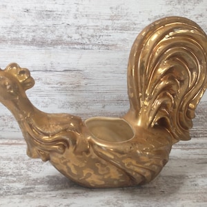 May include: A gold-colored ceramic rooster-shaped planter. The planter has a detailed design with a textured surface and a small opening for plants. The rooster is posed in a side profile, with its head and tail feathers prominently displayed.