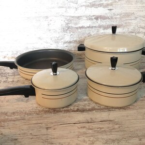 May include: A set of cream-colored enameled cookware featuring black handles and lids with black knobs. The pieces include a large lidded pot, two smaller lidded saucepans, and one open frying pan, all decorated with thin horizontal black and brown lines.