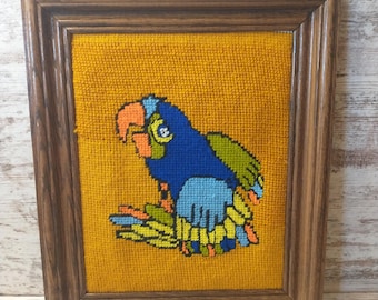 May include: Framed vintage needlepoint art featuring a colorful parrot. The parrot is primarily blue with accents of orange, green, and yellow, set against a mustard yellow background. The artwork is encased in a dark brown wooden frame.