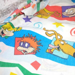 May include: White fabric with a colorful pattern featuring cartoon characters, stars, and geometric shapes. The design includes images of characters with orange, yellow, and green accents, set against blue, red, and green stripes.