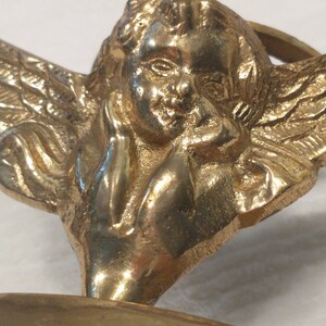 May include: A gold-toned metal cherub figurine with wings, holding its chin in its hand. The cherub is sitting on a round base.