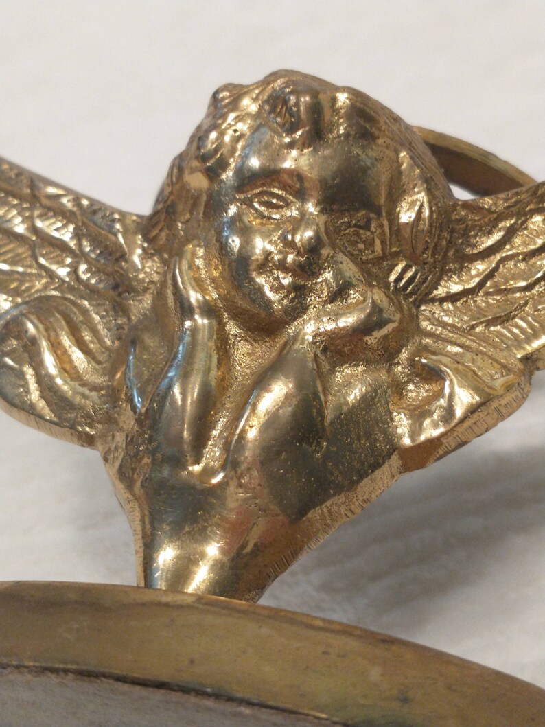 May include: A gold-toned metal cherub figurine with wings, holding its chin in its hand. The cherub is sitting on a round base.