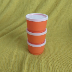 May include: Three orange plastic containers with pink lids stacked on top of each other. The containers are on a yellow textured surface.