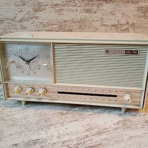 May include: Vintage General Electric AM/FM radio and clock combo in off-white. The clock face is on the left, with a black clock hands and white numbers. The radio has a horizontal speaker grill and tuning dial.