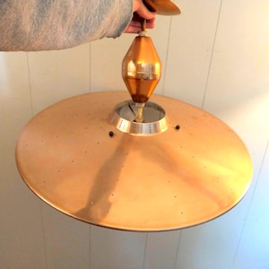 May include: A vintage mid-century modern pendant light fixture. The light has a large, round, copper-colored shade and a copper-toned, teardrop-shaped central element. The fixture is suspended from a thin rod.