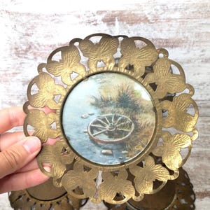 May include: Ornate brass-toned picture frame with a circular image of a water scene with a wagon wheel. The frame has a decorative butterfly and floral design around the edge. A vintage-style home decor item.