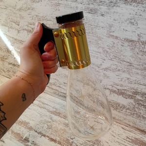 May include: A clear glass bottle with a gold-colored metal sleeve and a black handle. The bottle has a black cap and is held by a hand. The sleeve has a series of curved cutouts. The bottle has a unique shape.