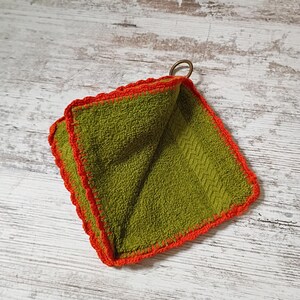 May include: A square olive green terry cloth washcloth featuring a vibrant orange crocheted border along all four edges. A small, circular bronze-colored metal ring is attached to one corner of the fabric, allowing for easy hanging or storage.