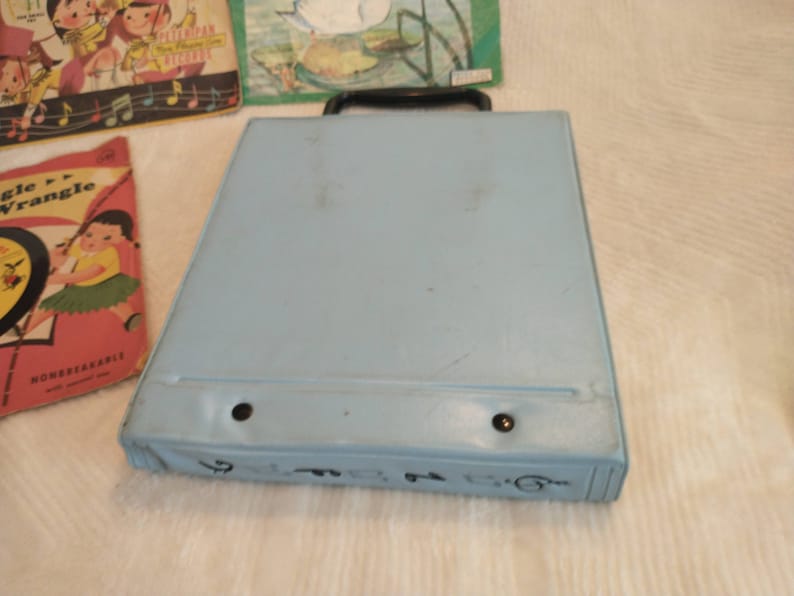 May include: A light blue record player case with a black handle. The case has two black dots on the front and a black line around the edge.