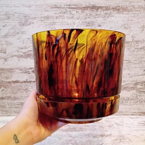 May include: A cylindrical glass planter featuring a translucent amber base color with swirling streaks of dark brown and black throughout the material. The vessel has a smooth, polished finish and a distinct horizontal ridge near the bottom edge.
