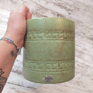 May include: A cylindrical, green ceramic planter with a textured, geometric pattern. The planter has a matte finish and a small, silver label that reads "Jaeger". The planter is held by a hand.