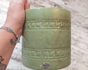 May include: A cylindrical, green ceramic planter with a textured, geometric pattern. The planter has a matte finish and a small, silver label that reads "Jaeger". The planter is held by a hand.