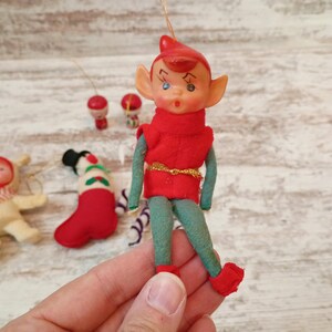 May include: A vintage Christmas elf ornament with a red hat and vest, green pants, and red shoes. The elf has a painted face and is holding a gold string. Other small ornaments are in the background.
