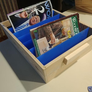 May include: A wooden magazine rack with blue dividers. The rack holds several magazines, including one with a person on the cover. The rack has a handle on the side and is sitting on a white surface.