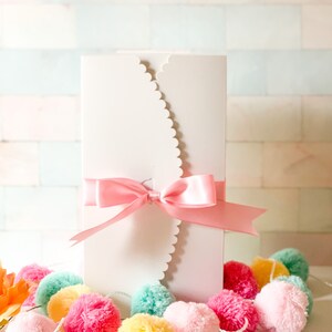 May include: White card with a scalloped edge and a pink satin ribbon tied in a bow. The card is surrounded by a garland of colorful pom-poms.