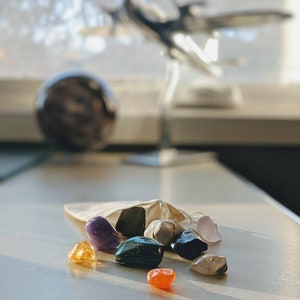 May include: A collection of colorful gemstones, including amethyst, rose quartz, and citrine, spilling from a small beige drawstring bag. A dark sphere and a metal airplane model are in the background.